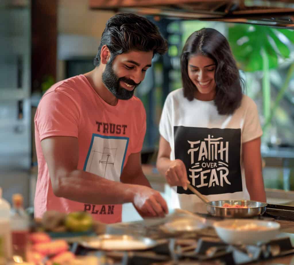 Man and woman cooking happily together. He is wearing a pink t-shirt with Trust his plan logo and she is wearing a white t-shirt with faith over fear on it